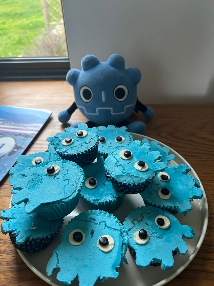 Our mascot plush sitting in front of a plate of cupcakes. They are frosted in bright blue, have googly eyes, and are shaped to mimick our logo.